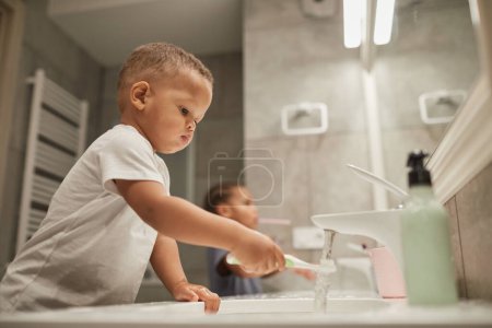 Photo for Side view portrait of two African American children in bathroom focus on little toddler boy brushing teeth - Royalty Free Image