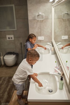 Téléchargez les photos : Portrait vertical latéral de deux enfants afro-américains dans la salle de bain accent sur le petit garçon brossant les dents - en image libre de droit