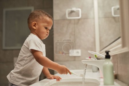 Side view portrait of African American toddler boy brushing teeth in bathroom, copy space