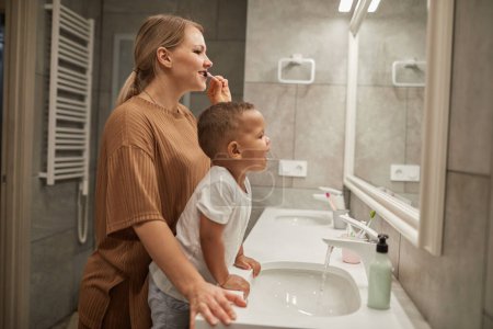 Téléchargez les photos : Vue de côté portrait de la jeune mère aidant mignon tout-petit garçon brossant les dents dans la salle de bain, espace de copie - en image libre de droit
