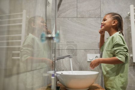 Foto de Retrato de vista lateral de niño negro feliz con pelo trenzado cepillando los dientes por espejo en el baño, espacio de copia - Imagen libre de derechos