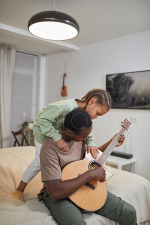 Téléchargez les photos : Portrait vertical de fille afro-américaine mignonne s'amuser avec papa jouer de la guitare dans la chambre à coucher - en image libre de droit