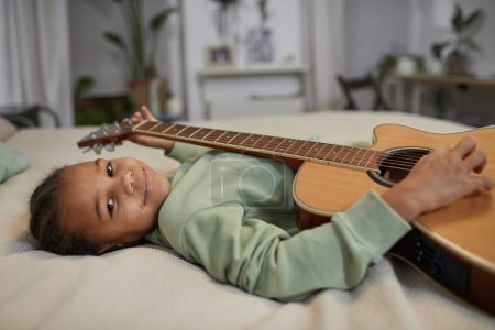 Foto de Retrato de linda chica negra sosteniendo la guitarra y mirando a la cámara mientras está acostado en la cama en casa, espacio de copia - Imagen libre de derechos