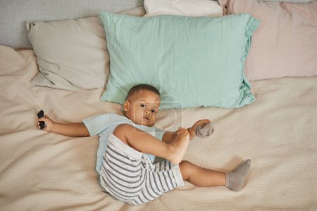 Photo for High angle view of cute black toddler laying on bed in pastel colored sheets and playing with socks looking at camera - Royalty Free Image