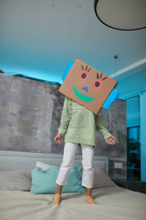 Photo for Full length portrait of unrecognizable child playing with cardboard box on head in blue colored bedroom - Royalty Free Image