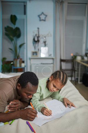 Foto de Retrato vertical de padre negro amoroso con hija dibujando juntos mientras que miente en la cama en el país, espacio de copia - Imagen libre de derechos