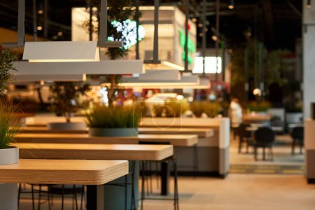 Téléchargez les photos : Image de fond de l'intérieur de la cour alimentaire dans le centre commercial avec des tables en bois en rangée éclairées par une lumière chaude, espace de copie - en image libre de droit