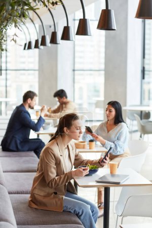 Téléchargez les photos : Portrait vertical de la jeune femme mangeant de la salade au food court et utilisant un smartphone, copier l'espace ci-dessus - en image libre de droit