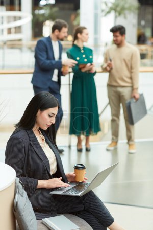 Téléchargez les photos : Portrait vertical d'une femme d'affaires du Moyen-Orient utilisant un ordinateur portable assis dans la salle d'attente d'un immeuble de bureaux moderne - en image libre de droit