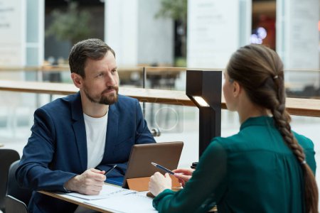 Téléchargez les photos : Portrait d'un homme d'affaires barbu écoutant une jeune femme lors d'une réunion au café avec des documents - en image libre de droit