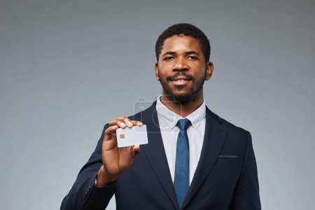 Foto de Retrato de hombre de negocios negro sonriente sosteniendo maqueta de tarjeta de crédito en blanco y mostrando a la cámara con fondo gris, espacio de copia - Imagen libre de derechos