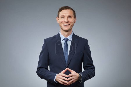 Photo for Waist up portrait of adult Caucasian man wearing business suit and smiling at camera while standing against grey background in studio, copy space - Royalty Free Image