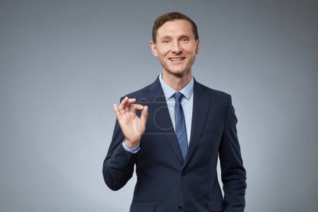 Photo for Waist up portrait of smiling Caucasian businessman showing OK sign while standing against grey background in studio, copy space - Royalty Free Image