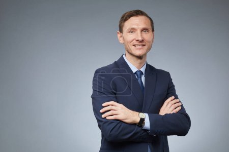 Photo for Waist up portrait of smiling Caucasian businessman standing with arms crossed and looking at camera against grey background, copy space - Royalty Free Image