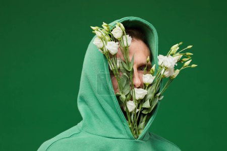 Foto de Retrato de hombre joven con rosas blancas de primavera en capucha de camisa sobre fondo verde vibrante, espacio de copia - Imagen libre de derechos