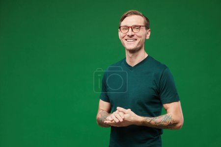 Téléchargez les photos : Portrait taille haute d'un homme tatoué portant une chemise verte et des lunettes sur fond vert vif en studio et souriant à la caméra, espace de copie - en image libre de droit