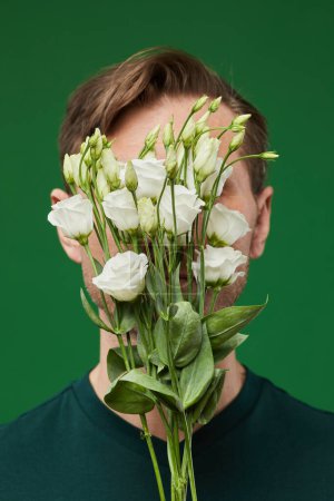 Foto de Retrato vertical del hombre cubriendo la cara con rosas blancas sobre un fondo verde vibrante - Imagen libre de derechos