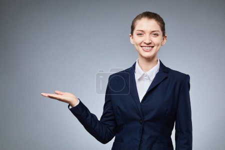 Foto de Retrato de cintura hacia arriba de una joven empresaria apuntando al espacio de copia y sonriendo a la cámara mientras está de pie sobre un fondo gris - Imagen libre de derechos