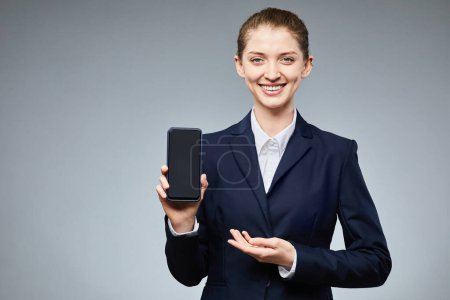 Téléchargez les photos : Portrait taille haute d'une femme d'affaires souriante tenant un smartphone avec écran blanc à la caméra sur fond gris, espace de copie - en image libre de droit