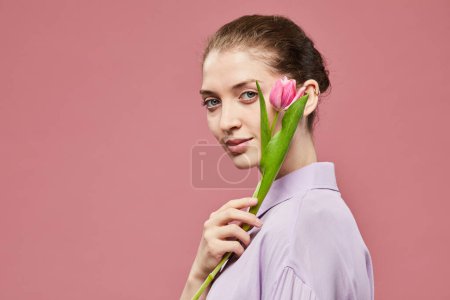 Téléchargez les photos : Portrait minimal d'une élégante jeune femme tenant une seule tulipe rose tout en se tenant debout sur un fond rose vif, espace de copie - en image libre de droit