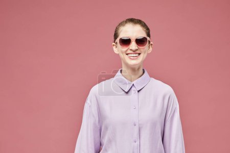 Foto de Cintura mínima hacia arriba retrato de la joven sonriente con gafas de sol y traje rosa sobre fondo rosa vibrante en el estudio, espacio de copia - Imagen libre de derechos