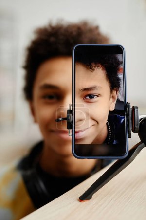 Téléchargez les photos : Portrait divisé vertical d'une adolescente noire souriante filmant une vidéo pour les médias sociaux, mise au point sur l'écran du smartphone sur le trépied - en image libre de droit