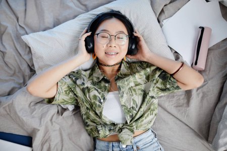 Téléchargez les photos : Vue du dessus portrait de l'adolescente asiatique portant des écouteurs et couché sur le lit souriant à la caméra avec des bretelles - en image libre de droit