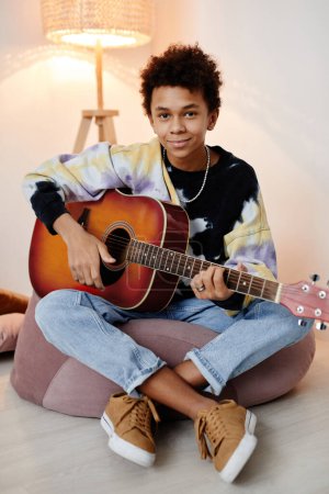 Photo for Vertical full length portrait of black teenage boy playing guitar and smiling at camera while sitting on bean bag - Royalty Free Image