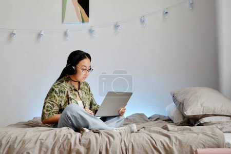 Téléchargez les photos : Minimal pleine longueur portrait de l'adolescente asiatique à l'aide d'un ordinateur portable avec écouteurs tout en étant assis sur le lit jambes croisées, espace de copie - en image libre de droit