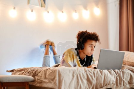 Foto de Retrato de niño afroamericano adolescente usando computadora portátil mientras está acostado en la cama en el interior de la habitación mínima, espacio de copia - Imagen libre de derechos