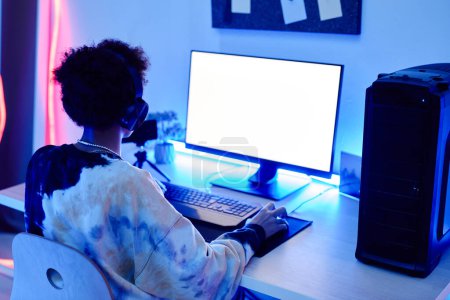 Téléchargez les photos : Vue arrière du garçon adolescent jouant au jeu vidéo sur PC et en streaming en direct dans la lumière bleue néon avec maquette d'écran blanc, espace de copie - en image libre de droit