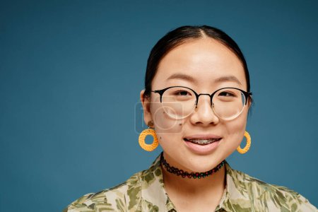 Téléchargez les photos : Portrait minimal d'une adolescente asiatique portant des bretelles et regardant la caméra sur fond bleu, espace de copie - en image libre de droit