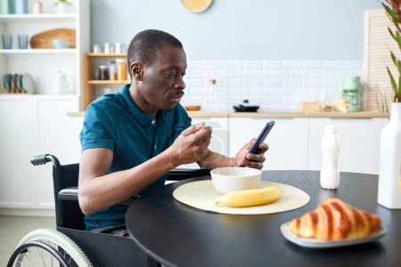 Foto de Retrato de hombre adulto negro con discapacidad desayunando en la cocina casera y usando teléfono, espacio para copiar - Imagen libre de derechos