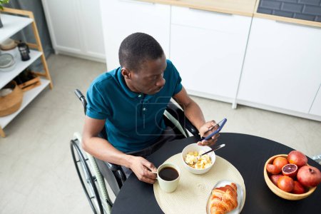 Téléchargez les photos : Vue du dessus de l'homme noir adulte handicapé prenant le petit déjeuner dans la cuisine de la maison et utilisant le téléphone, l'espace de copie - en image libre de droit