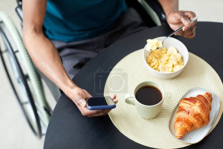 Téléchargez les photos : Vue du dessus d'un homme méconnaissable avec un handicap prenant le petit déjeuner à la maison et utilisant le téléphone, l'espace de copie - en image libre de droit