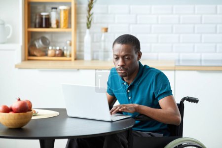 Téléchargez les photos : Portrait d'un homme afro-américain adulte handicapé utilisant un ordinateur portable à la maison dans un intérieur de cuisine minimal, espace de copie - en image libre de droit