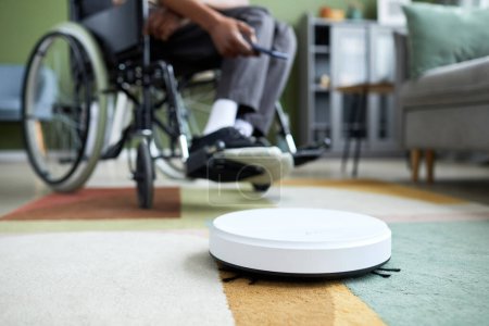 Foto de Primer plano de la aspiradora robot en la alfombra en el interior del hogar con el hombre usando silla de ruedas en el fondo, espacio de copia - Imagen libre de derechos
