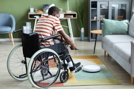 Photo for Full length portrait of modern black man with disability using robot vacuum cleaner and smart home technology, copy space - Royalty Free Image