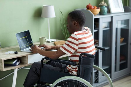 Photo for Side view portrait of black man in wheelchair using laptop and writing code while working from home - Royalty Free Image