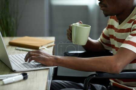 Téléchargez les photos : Vue de côté gros plan de l'homme afro-américain en fauteuil roulant en utilisant un ordinateur portable tout en travaillant à la maison et en buvant du café - en image libre de droit