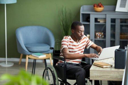 Foto de Retrato de hombre afroamericano con discapacidad utilizando el ordenador portátil mientras trabaja desde casa contra la pared verde, espacio de copia - Imagen libre de derechos