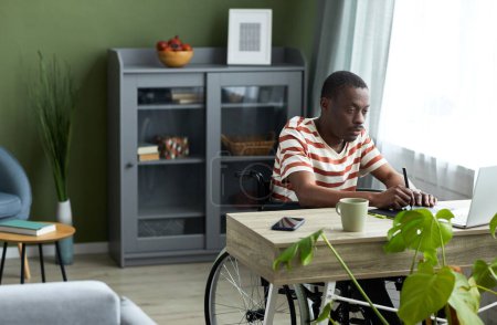 Téléchargez les photos : Portrait d'un homme noir adulte en fauteuil roulant utilisant une tablette de stylo tout en travaillant avec un ordinateur à la maison, espace de copie - en image libre de droit