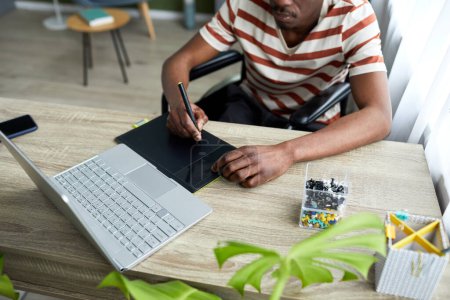 Téléchargez les photos : Gros plan de l'homme afro-américain en fauteuil roulant utilisant une tablette de stylo tout en travaillant avec un ordinateur à la maison, espace de copie - en image libre de droit