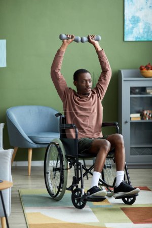 Foto de Retrato vertical de cuerpo entero del hombre afroamericano con discapacidad haciendo ejercicio usando pesas en casa - Imagen libre de derechos