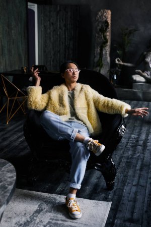 Photo for Vertical full length portrait of Asian man in extravagant outfit lounging in leather armchair at dark studio - Royalty Free Image
