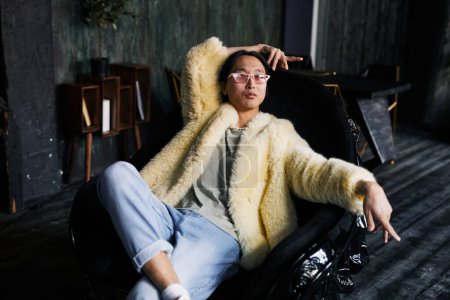 Téléchargez les photos : Portrait d'homme asiatique en tenue extravagante avec manteau en fourrure néon se prélasser dans un fauteuil en cuir au studio sombre - en image libre de droit