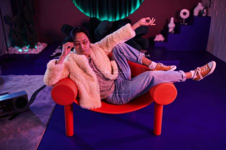 Photo for Full length portrait of Asian man wearing extravagant outfit lying across chair in pink neon light - Royalty Free Image