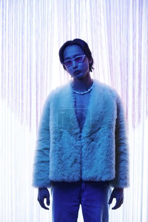 Photo for Vertical portrait of extravagant Asian man wearing fur coat at nightclub or party in blue light - Royalty Free Image