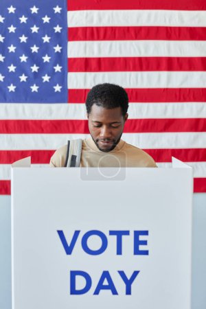 Téléchargez les photos : Portrait vertical du jeune homme noir debout dans l'isoloir le jour du scrutin et faisant des choix politiques - en image libre de droit