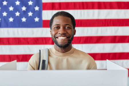 Téléchargez les photos : Portrait d'un homme noir souriant debout dans une isoloir et souriant à la caméra contre le drapeau américain - en image libre de droit